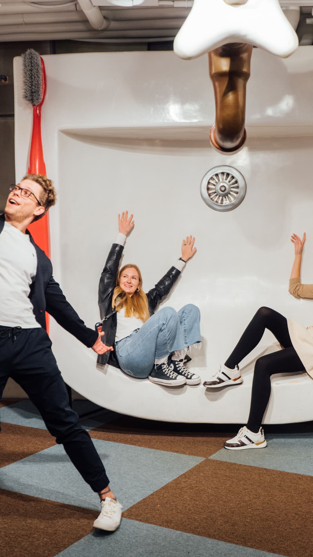 Two friends lounging inside a giant bathroom sink while a man takes a selfie, surrounded by an enormous toothbrush, toothpaste tube and plunger at The Giant's House Barcelona