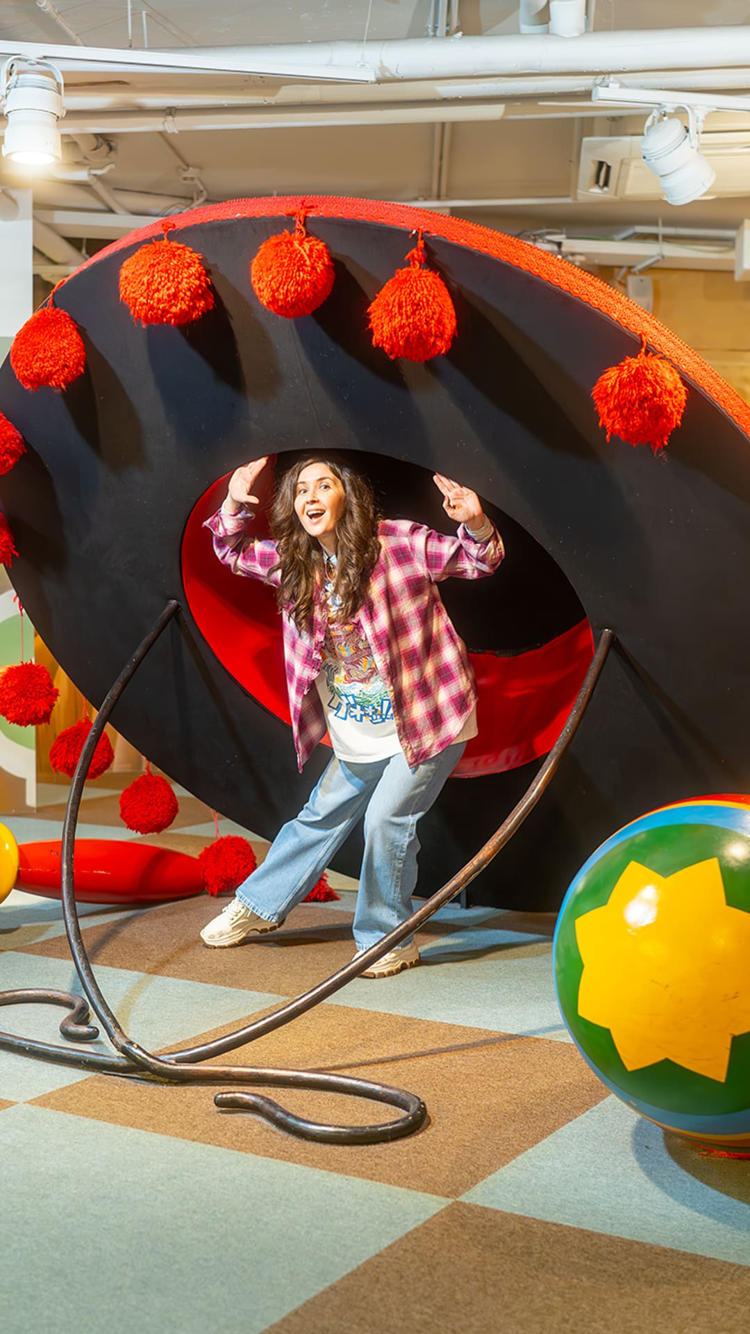 Woman striking a pose inside a giant black tambourine ringed with red pom-poms, beside oversized painted spinning tops at The Giant's House Barcelona