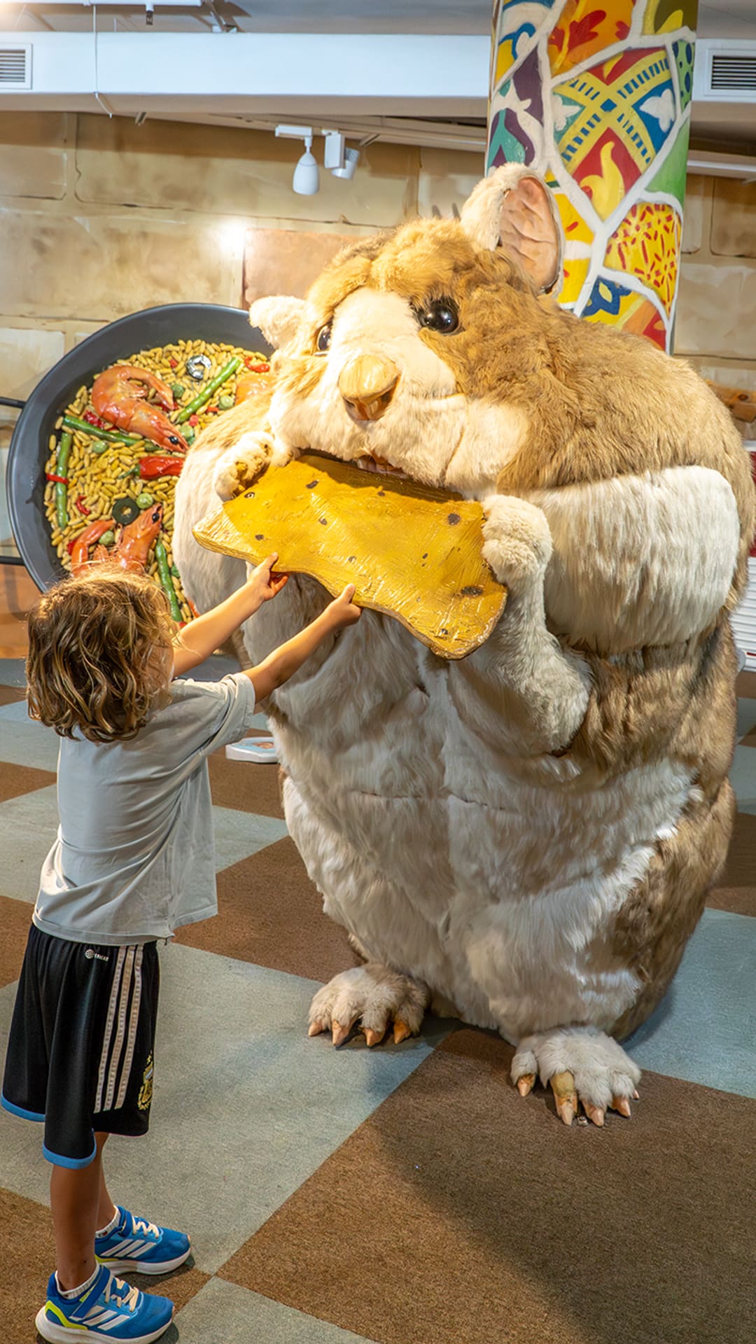 Child reaching up to feed a giant wedge of cheese to an enormous fluffy hamster, with a huge paella pan and oversized soup can nearby at The Giant's House Barcelona